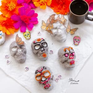 square crop of 6 skull cakes decorated for Day of the Dead with a cup of coffee and flowers in the background.