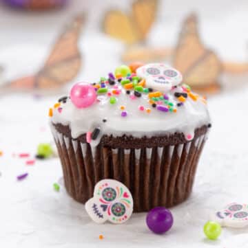 A chocolate cupcake with white icing decorated with sprinkles and monarch butterflies in the background.