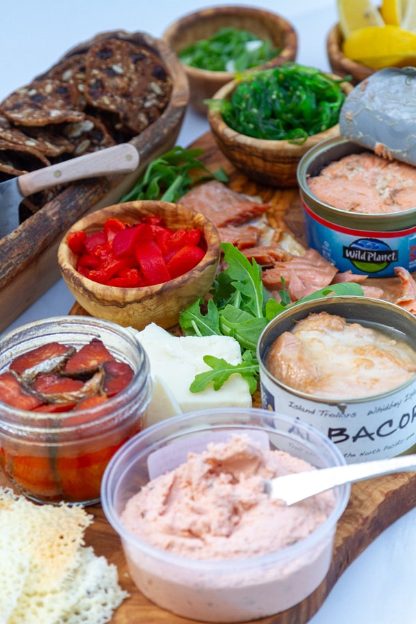 an olive wood board filled with smoked salmon pate, tinned salmon and tuna, red peppers, arugula, cheese, frico, seaweed salad, and served with crackers on the side.