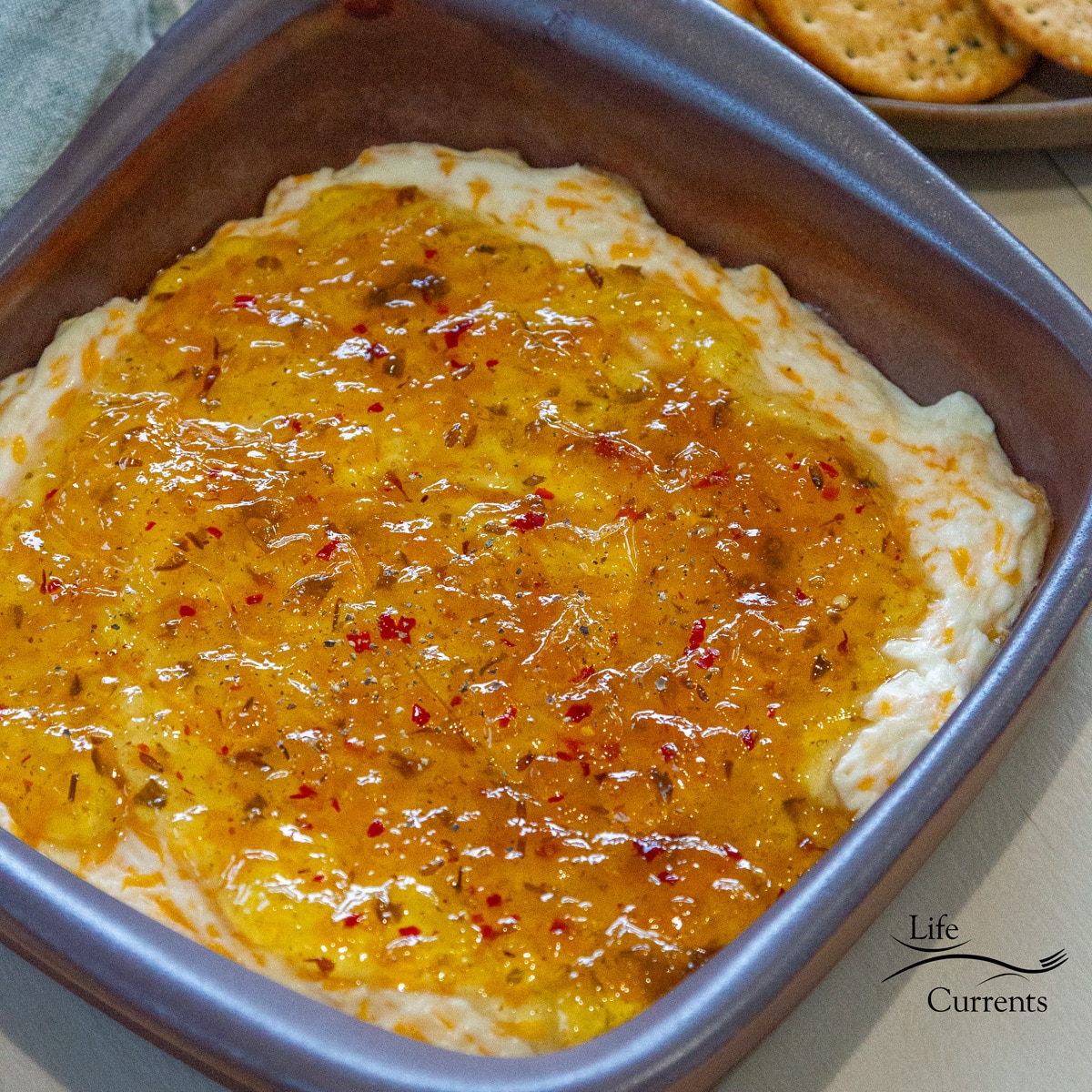 square crop looking down into a baking dish filled with pepper jelly topped cheese dip with crackers on the right.