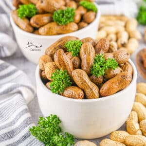 two white bowls filled with cooked peanuts in the shell garnished with parsley and there's a striped towel on the left.