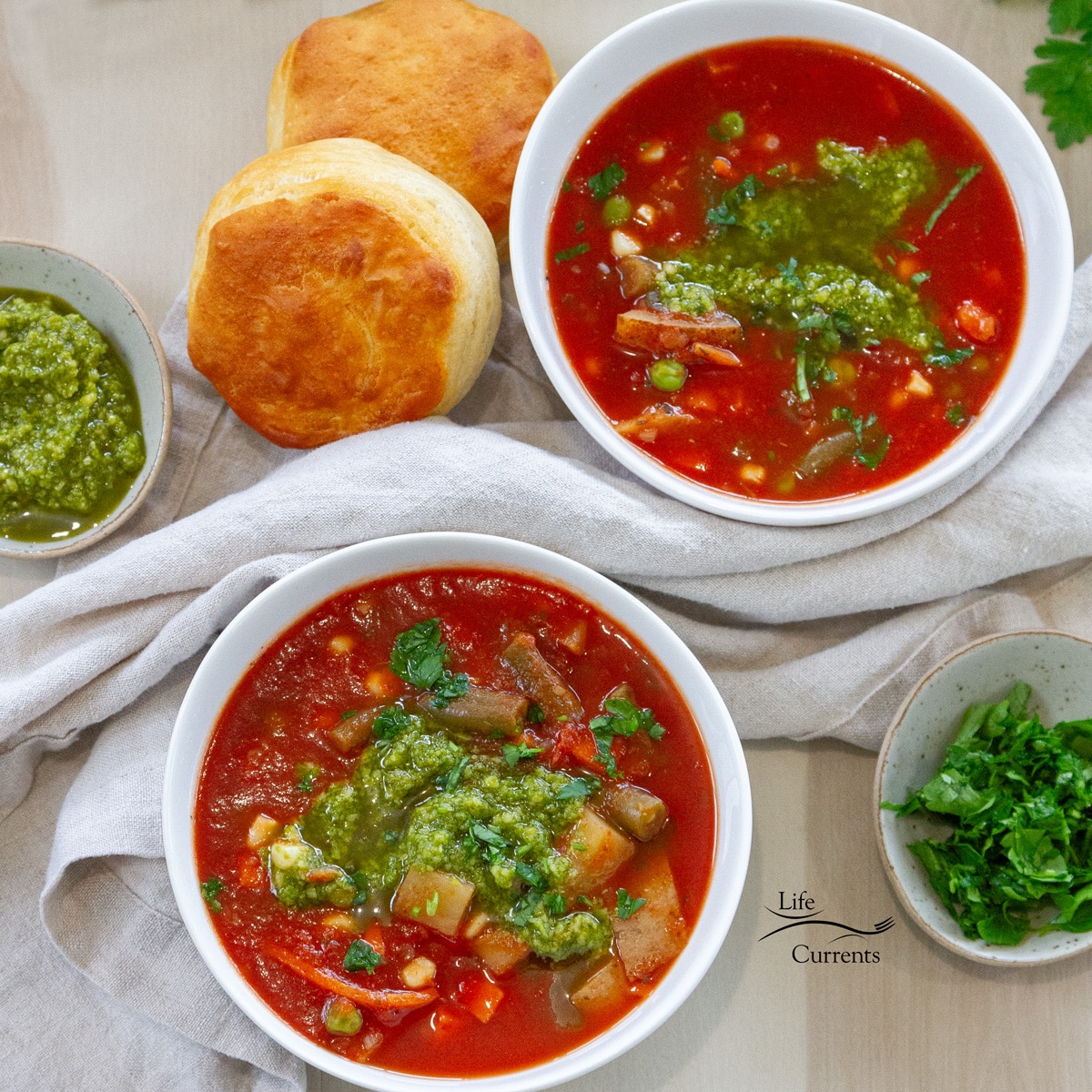 square crop of two white bowls filled with soup, two biscuits to the upper left and two bowls of green garnishes.