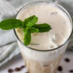 square crop of a glass of coffee topped with cream and a mint sprig.