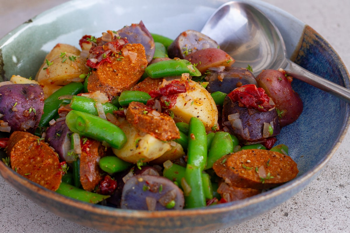 a bowl filled with sausage potatoes and snap peas and a serving spoon in the back.