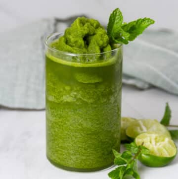 square crop of a matcha mojito in a glass garnished with fresh mint and some squeezed limes next to the glass.