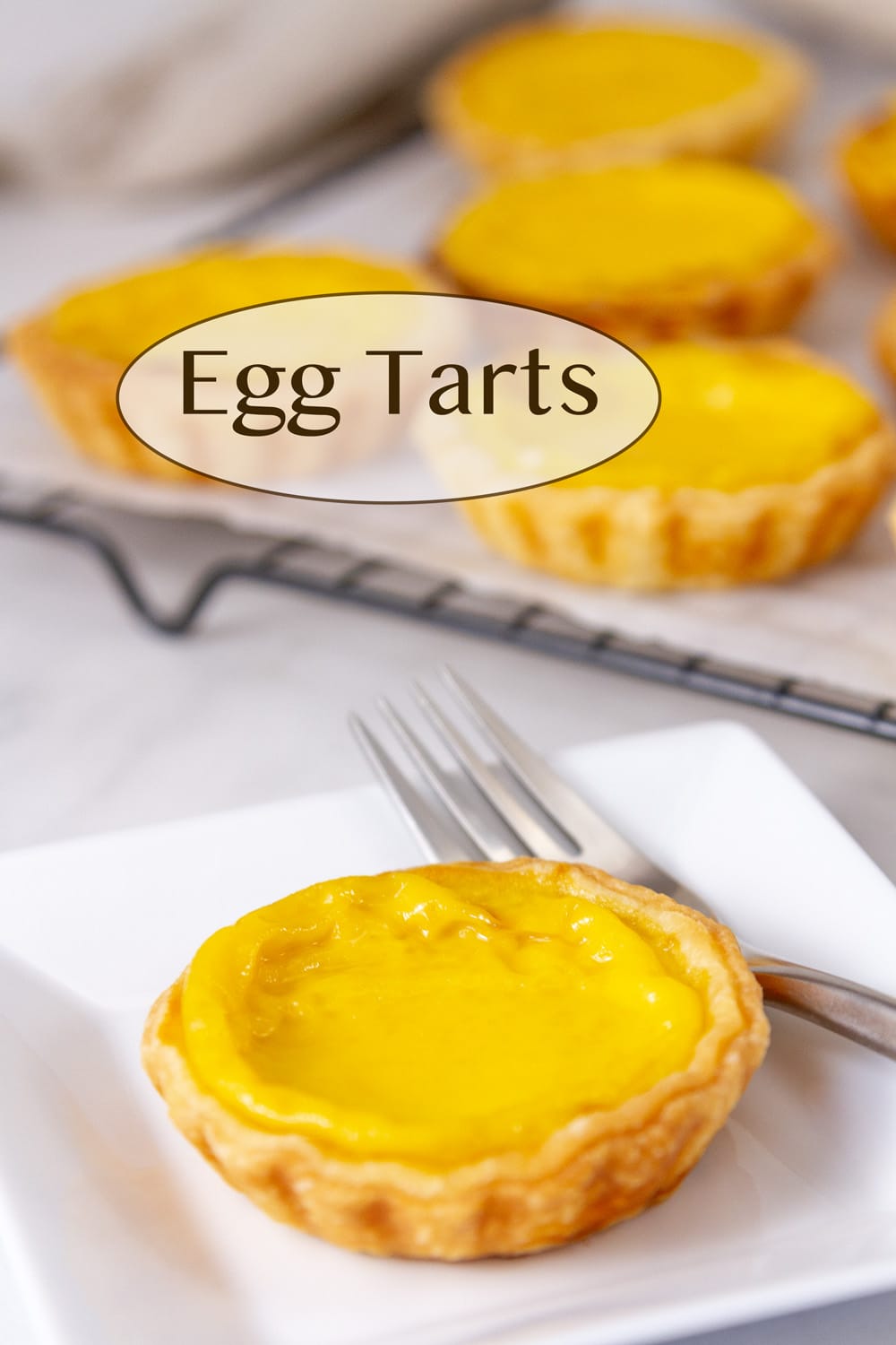 an egg tart on a white plate with a fork in front of some tarts cooling on a wire rack.