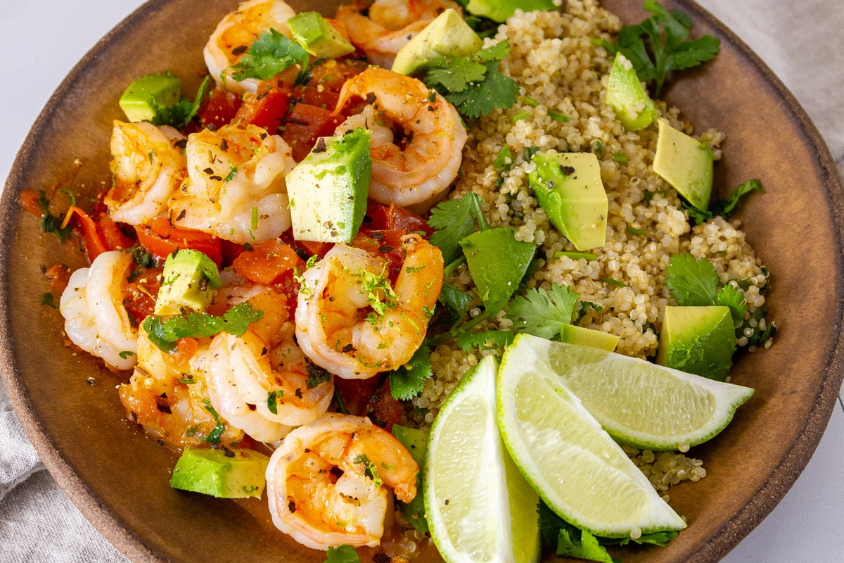 a brown bowl filled with shrimp, tomatoes, quinoa, avocado, and lime wedges.