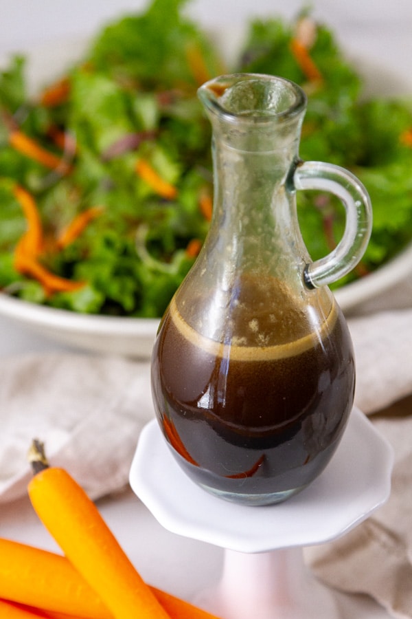 a bottle of salad dressing on a small white pedestal in front of a large bowl of salad.