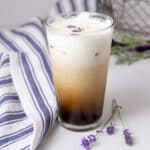 square crop of a latte topped with cold foam in a glass with a towel on the side and lavender flowers next to the glass.