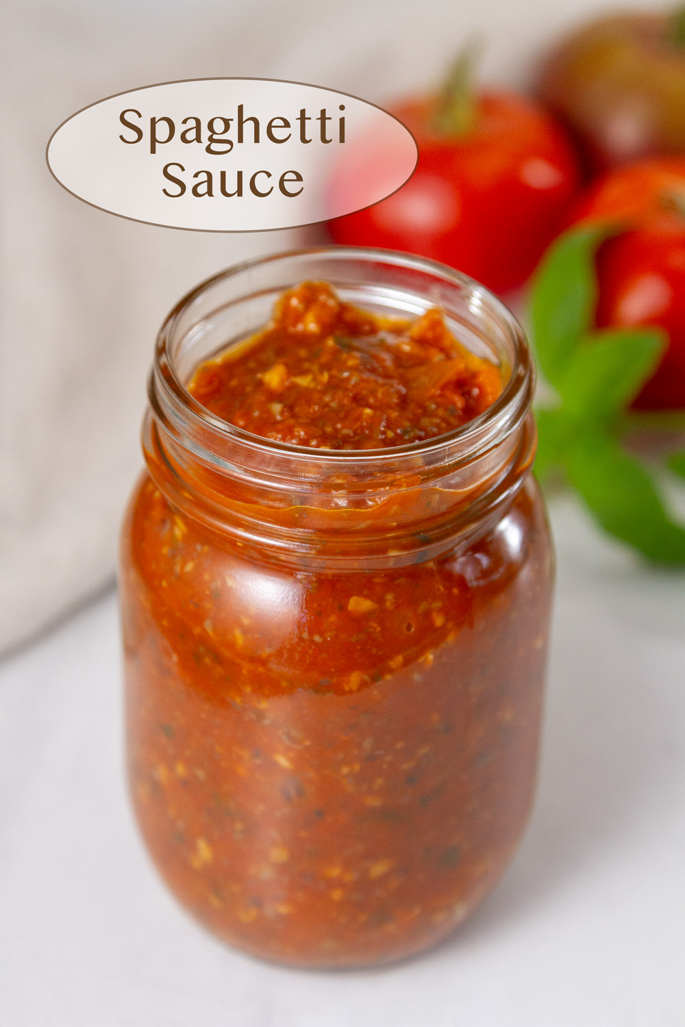 a glass jar filled with red spaghetti sauce and some tomatoes and basil in the background.