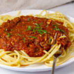 square crop of bucatini spaghetti pasta on a white plate covered with red spaghetti sauce and a fork in the pasta.