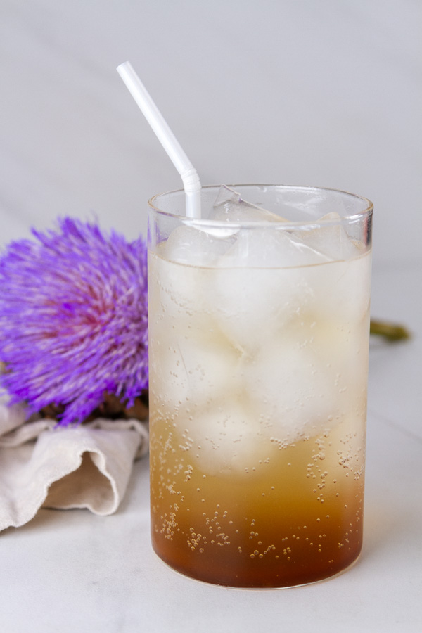 a cardamom mocktail drink over ice with a straw in front of an artichoke flower.