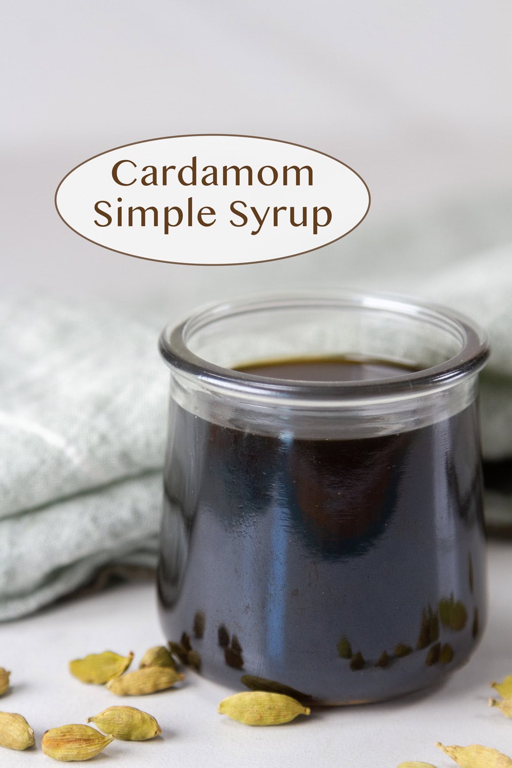 cardamom simple syrup in a small glass jar with cardamom pods around it and a green cloth napkin behind it.