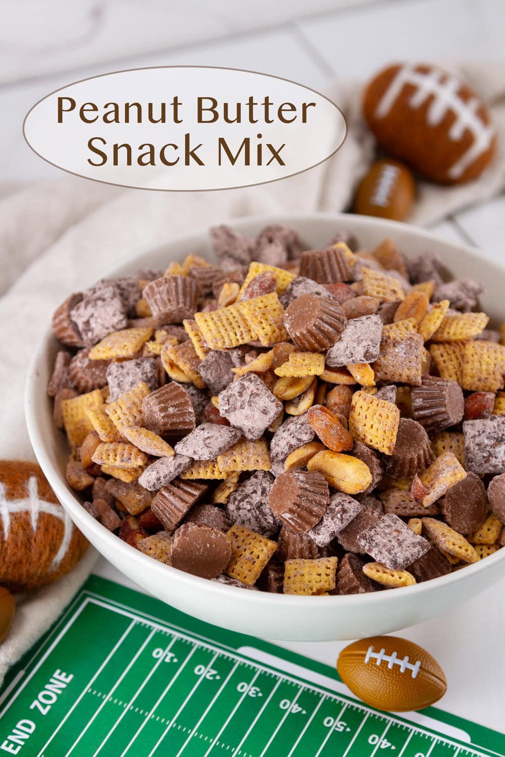 A white bowl filled with a sweet snack mix of cereal and peanut butter cups with some footballs around the bowl and a green football field mat.