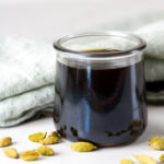 square crop of a glass jar filled with syrup and cardamom pods around it.