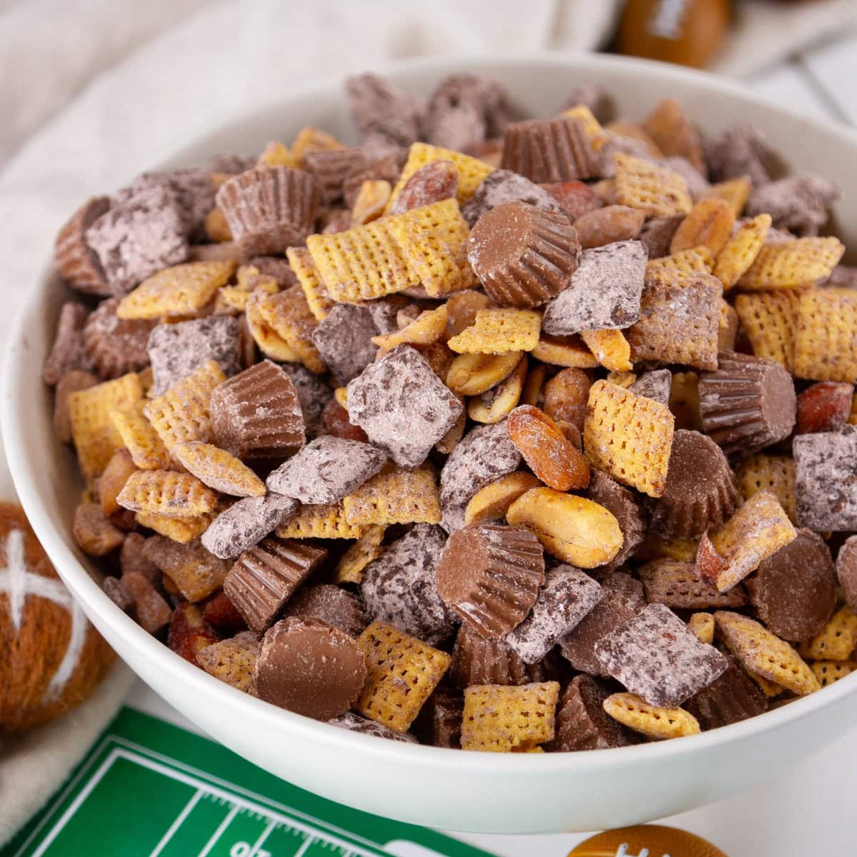 square crop of a white bowl filled with peanut butter snack mix and some football decorations around the bowl.