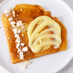 square crop of a crepe on a white plate topped with lemon curd and crumbled meringue.