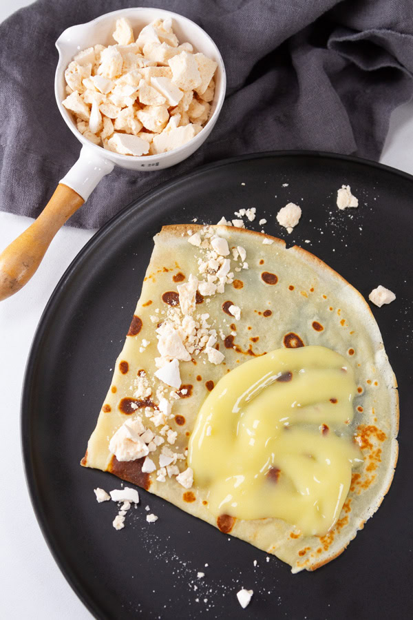 a lemon meringue crepe on a black plate with a dish full of crumbled meringues to the upper left.