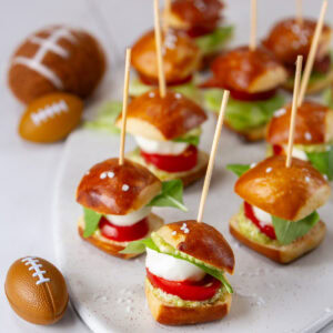 square crop of mini pretzel bites sandwiches on a white serving tray with some small footballs in the background.