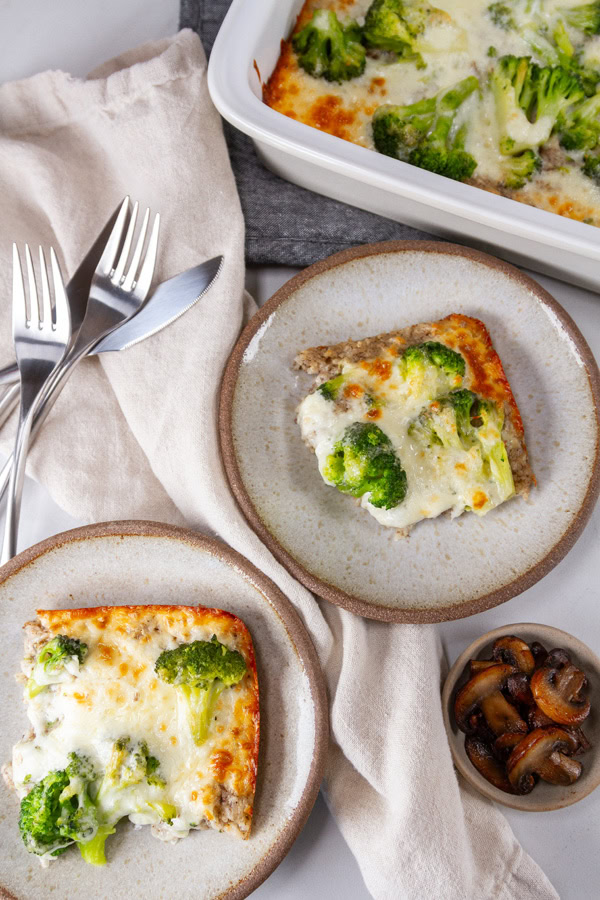 two servings of casserole on small plates with forks, knives, and a bowl of mushrooms.