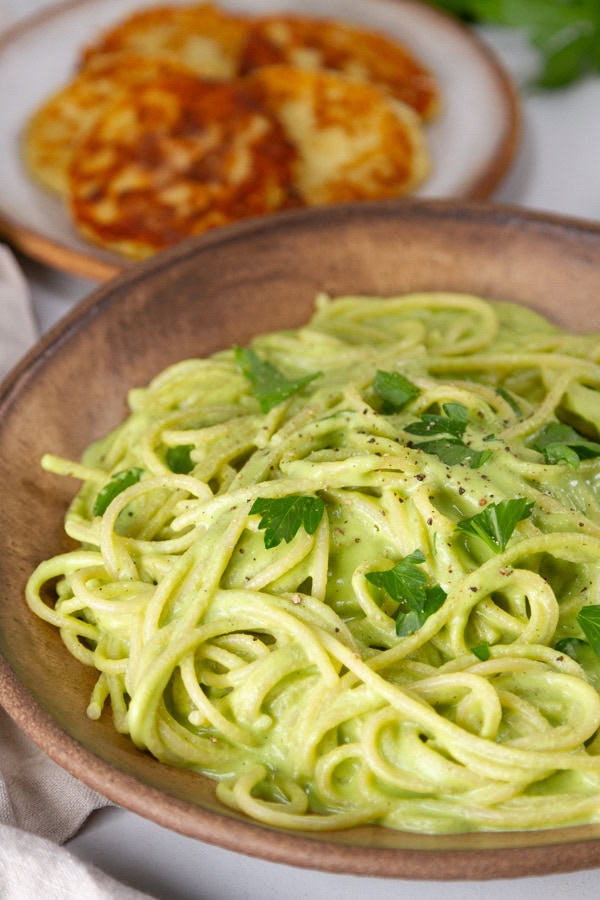 pasta in green sauce in a brown bowl with tortitas de papa in the background.