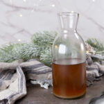 square crop of a glass jar filled with ginger simple syrup in front of a cloth and some greenery.