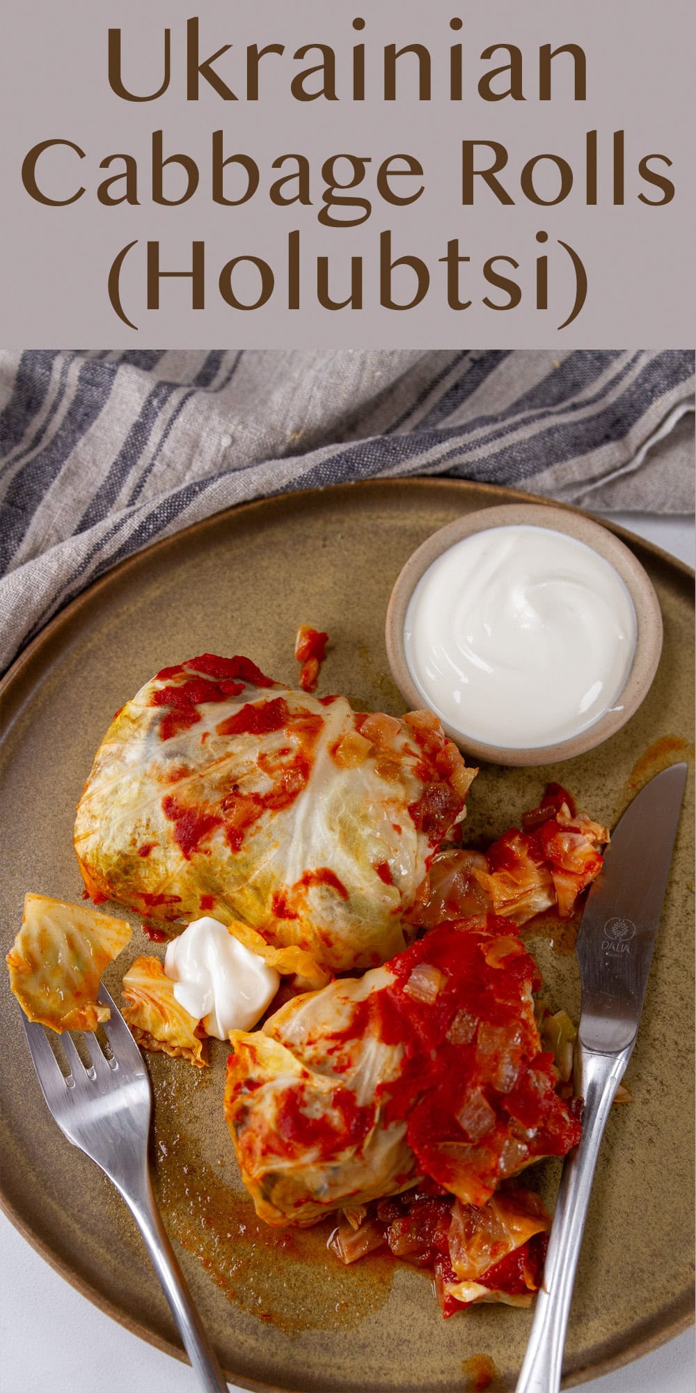 looking down on a plate with two cabbage rolls and a small dish of sour cream there's a fork and a knife as well.