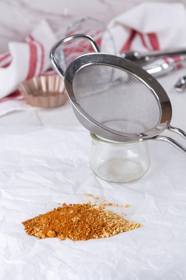spices on a piece of parchment paper with a glass jar and a strainer next to it.
