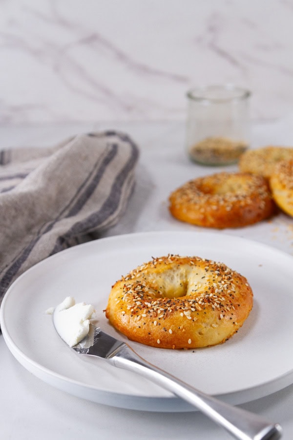 a bagel on a white plate with a cream cheese covered knife and some bagels and a cloth napkin in the background.