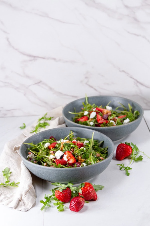 two blue bowls filled with colorful salad and some strawberries around them.