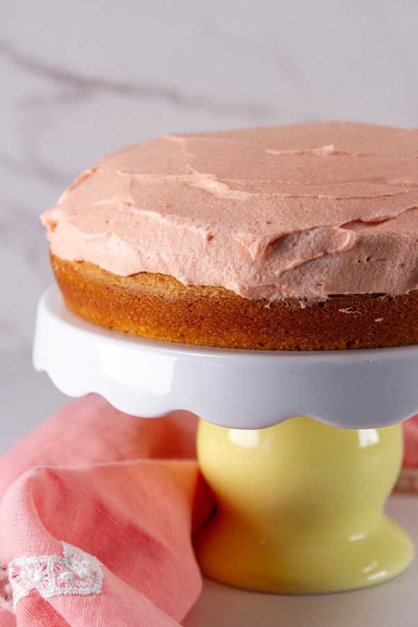 frosted cake on a cake stand with a pink cloth napkin in the background.