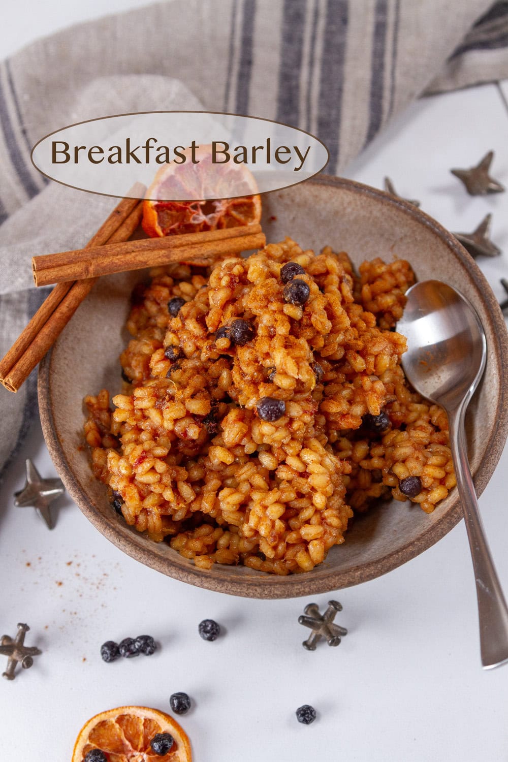 a bowl filled with cooked barley and fruit with a spoon on the right and cinnamon sticks and other spices around the bowl.