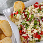 square crop of a tray of 7 Layer Mediterranean Dip next to a plate of naan and a naan in the dip.
