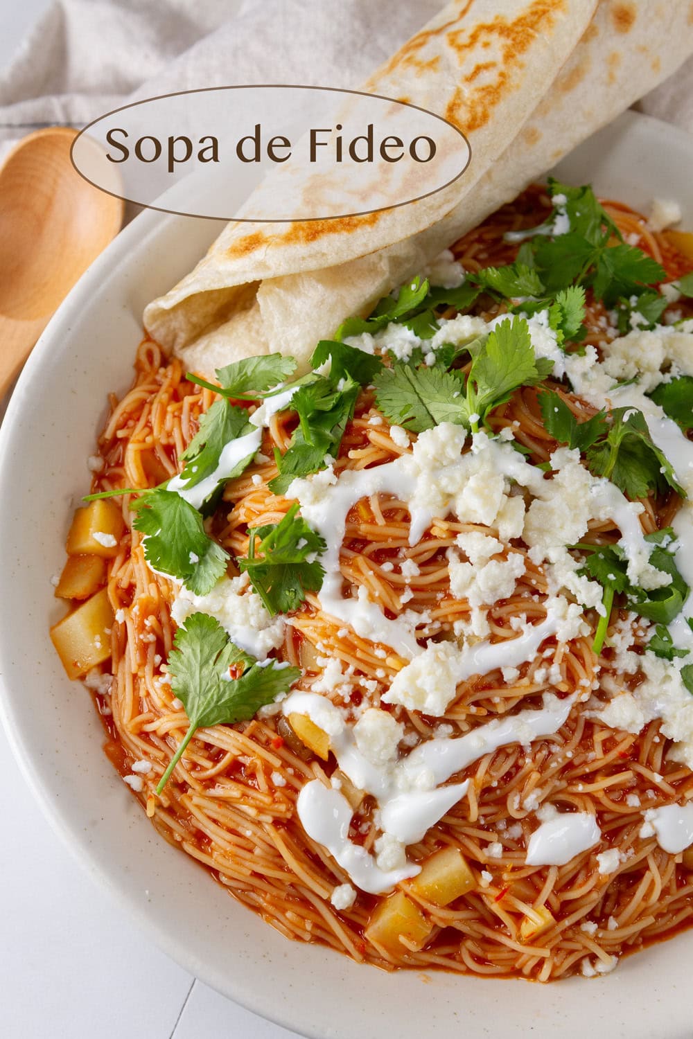 a bowl of soup with fideo pasta and tomato broth topped with cilantro queso fresco and sour cream and served with a tortilla.