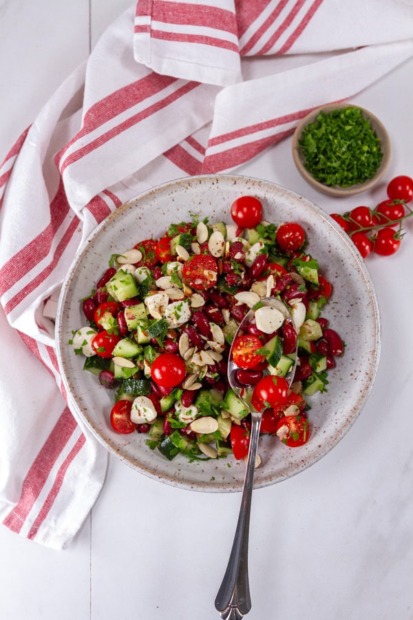 a bowl of salad with a serving spoon next to a red striped cloth, cherry tomatoes, and a bowl of chopped parsley.