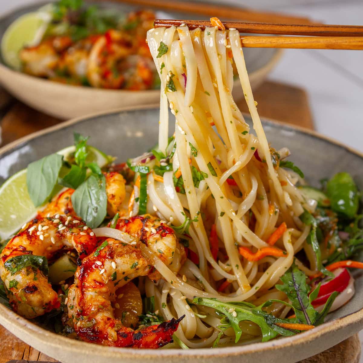 square crop of chopsticks pulling up noodles out of a bowl with shrimp and salad in it.