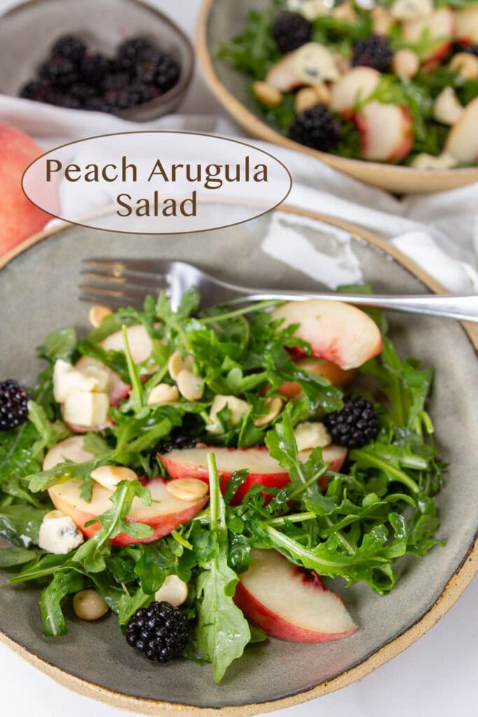 a bowl of arugula salad with peaches, blackberries, cheese, and nuts and a fork in the bowl.