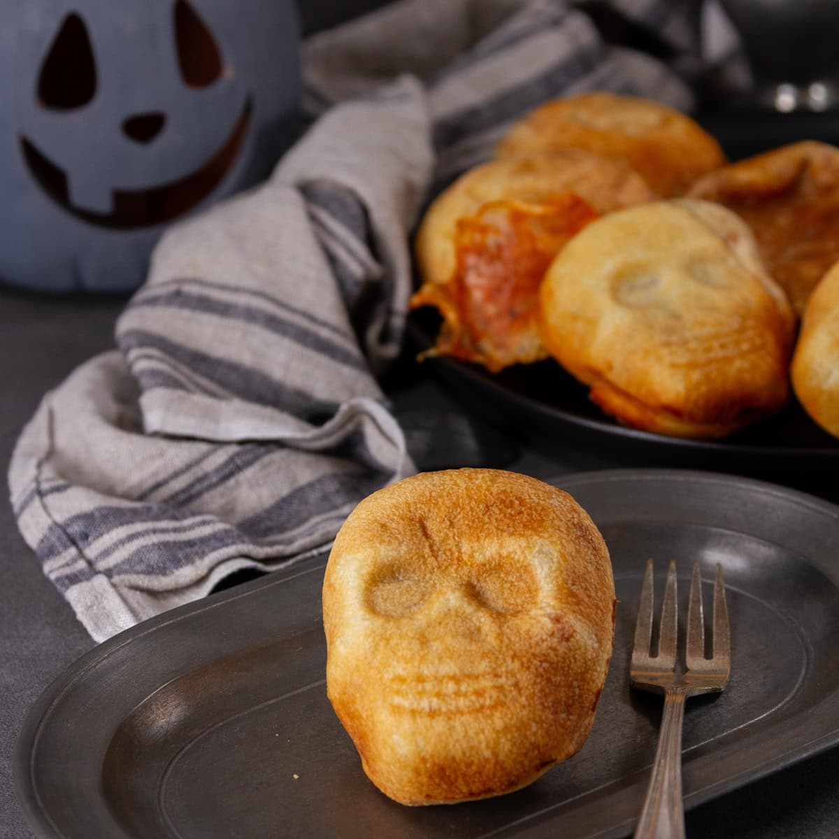 square crop of pizza calzones in the shape of skulls for Halloween with a fork on the plate and a jack o' lantern behind.