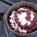 square crop of a bowl of black rice pudding topped with coconut milk and a spoon to the left.