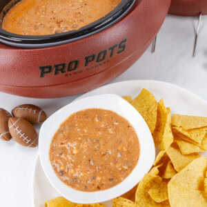 square crop of chili cheese dip with chips in front of a football shaped slow cooker.