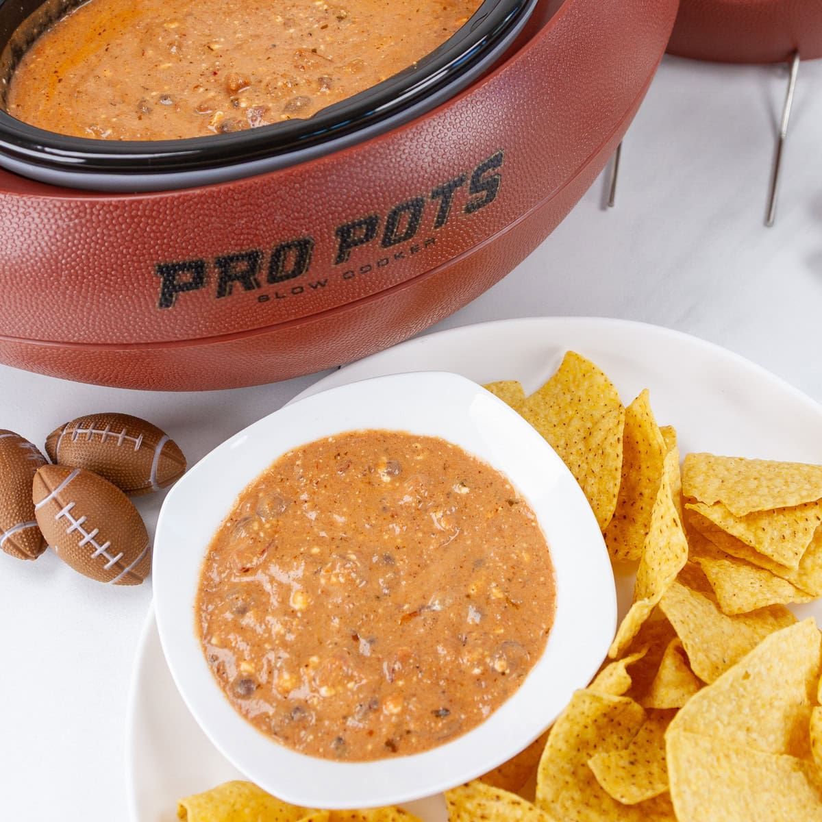 square crop of chili cheese dip with chips in front of a football shaped slow cooker.