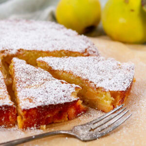 square crop of a cake that's been sliced with a fork and some fruit behind.