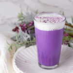 square crop of an iced ube latte with cold foam in front of some flowers.