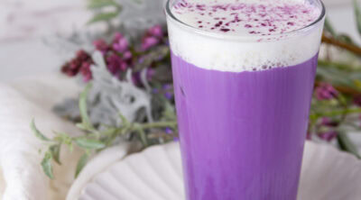 square crop of an iced ube latte with cold foam in front of some flowers.