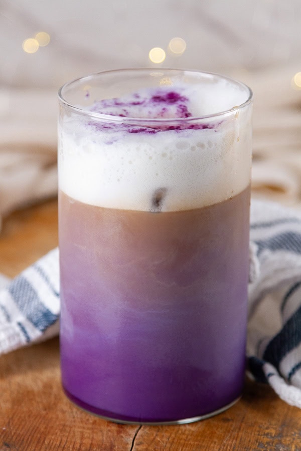 a glass with ube latte inside on a wooden board with a blue striped towel.