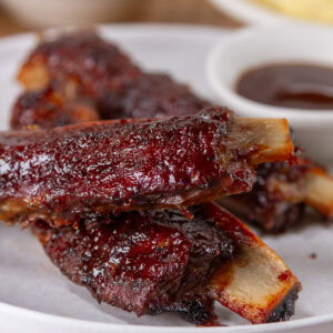 square crop of ribs piled on a white plate with a small dish of BBQ sauce in the background.