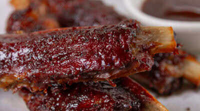 square crop of ribs piled on a white plate with a small dish of BBQ sauce in the background.