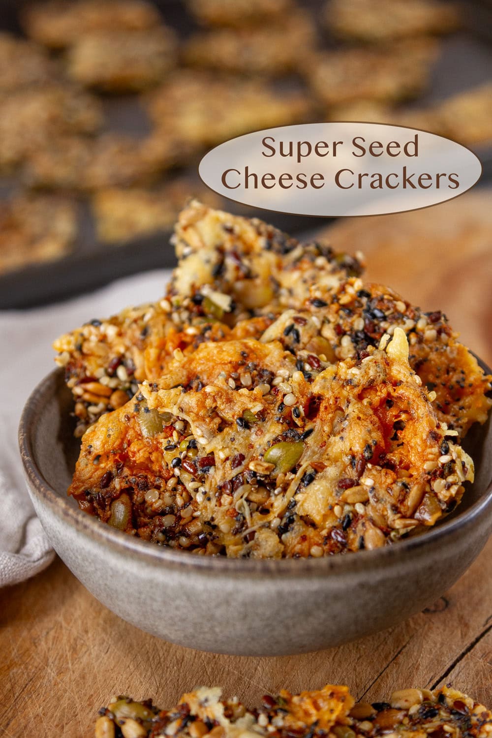 a small bowl filled with seed and cheese crackers on a wooden background with a tray of crackers behind the bowl.