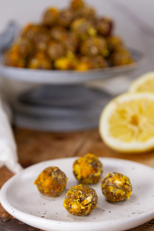 4 energy bites on a small white plate in front of the serving plate full of them and some lemon slices in the background.