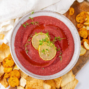 square crop of pink colored beet hummus with slices of lemon garnish in a white bowl with crackers and pita chips around.